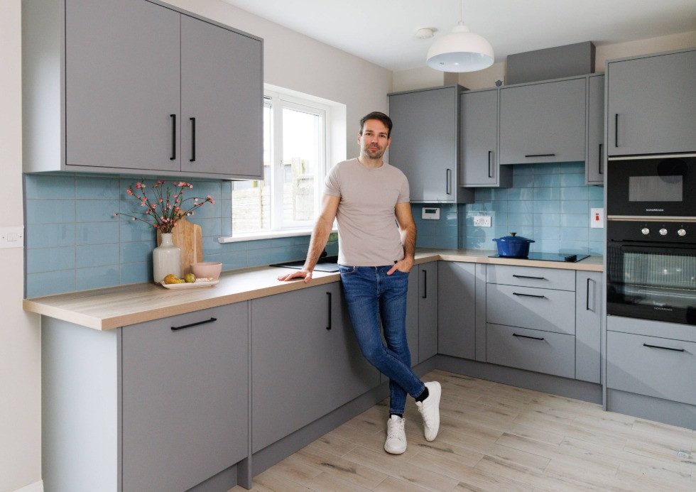Man in corner of modern light grey kitcehn with wooden worktop, floor, black handles and appliances